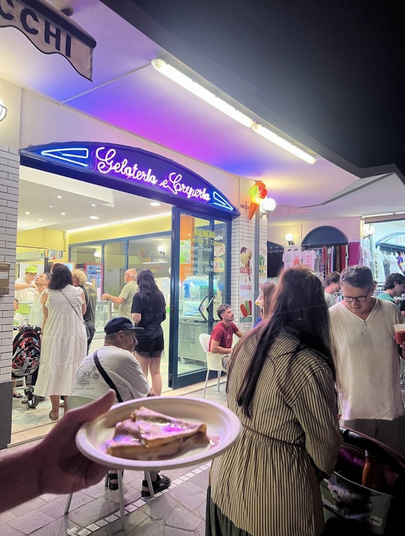 L'insegna al neon della Gelateria Creperia Dolomiti di sera, con clienti in fila